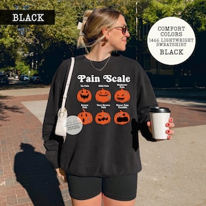 May include: A black long-sleeve sweatshirt featuring a graphic of six orange jack-o'-lanterns arranged in a grid. The text reads Pain Scale, with labels for No Pain, Mild Pain, Moderate Pain, Severe Pain, Very Severe Pain, and Worst Pain Possible.