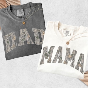 Puede incluir: Dos camisetas, una gris oscuro y otra blanquecina, cada una con una palabra en estampado de camuflaje: "DAD" y "MAMA". Ambas camisetas tienen un collar dorado con un colgante. Las camisetas est&aacute;n dobladas y expuestas sobre un fondo claro.