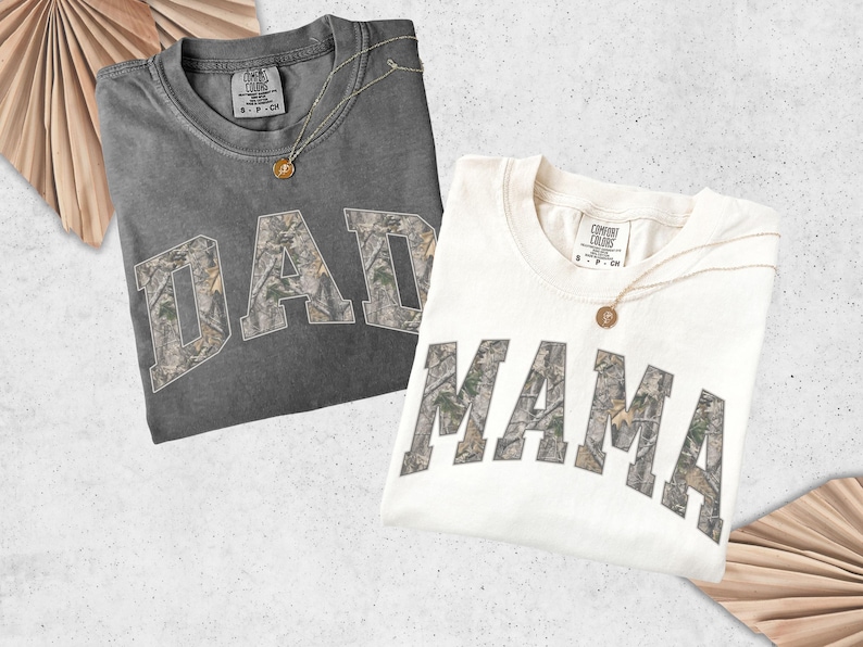 Puede incluir: Dos camisetas, una gris oscuro y otra blanquecina, cada una con una palabra en estampado de camuflaje: "DAD" y "MAMA". Ambas camisetas tienen un collar dorado con un colgante. Las camisetas est&aacute;n dobladas y expuestas sobre un fondo claro.