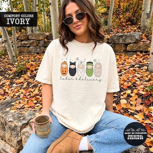 K&ouml;nnte beinhalten: Ein elfenbeinfarbenes Kurzarm-T-Shirt mit einer Grafik von f&uuml;nf in Windeln gewickelten Babys, die als K&uuml;rbis, Fledermaus, Geist, Frankensteins Monster und Mumie verkleidet sind, mit dem Text "labor & delivery" in schwarzer Schreibschrift darunter.
