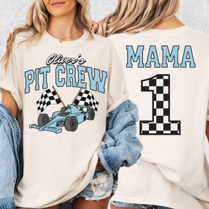 May include: Ivory-colored t-shirt with "Oliver's PIT CREW" in light blue, featuring a race car and checkered flags. The back displays "MAMA" and a checkered number "1" in black and white.