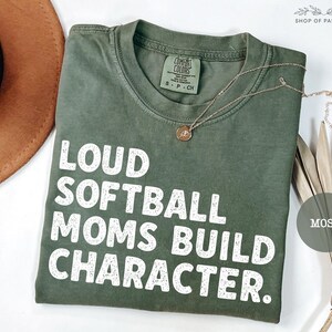 May include: A moss green t-shirt with the text "LOUD SOFTBALL MOMS BUILD CHARACTER" in white block letters. A gold necklace with a pendant rests on the shirt. The shirt is folded and displayed with a brown hat and dried plant.
