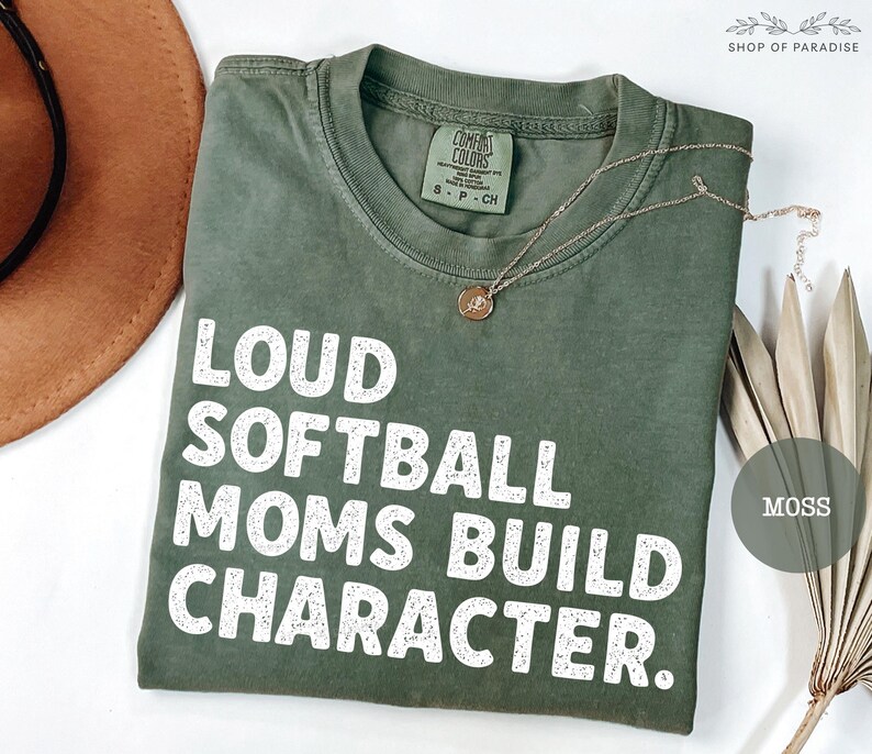 May include: A moss green t-shirt with the text "LOUD SOFTBALL MOMS BUILD CHARACTER" in white block letters. A gold necklace with a pendant rests on the shirt. The shirt is folded and displayed with a brown hat and dried plant.