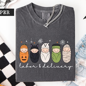 K&ouml;nnte beinhalten: Ein anthrazitfarbenes T-Shirt mit einer Grafik von f&uuml;nf in Windeln gewickelten Babys, die als K&uuml;rbis, Fledermaus, Geist, Frankensteins Monster und Mumie verkleidet sind. Darunter steht der Text labor & delivery in wei&szlig;er Schreibschrift.