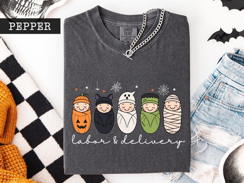 K&ouml;nnte beinhalten: Ein anthrazitfarbenes T-Shirt mit einer Grafik von f&uuml;nf in Windeln gewickelten Babys, die als K&uuml;rbis, Fledermaus, Geist, Frankensteins Monster und Mumie verkleidet sind. Darunter steht der Text labor & delivery in wei&szlig;er Schreibschrift.