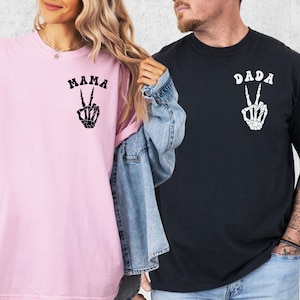 May include: Two short-sleeved t-shirts, one pink and one black. The pink shirt has the word "MAMA" above a skeleton hand making a peace sign. The black shirt has the word "DADA" above a skeleton hand peace sign.