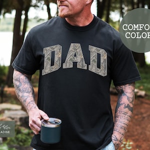 Puede incluir: Camiseta negra con la palabra "DAD" en un dise&ntilde;o de camuflaje en el pecho. La camiseta es de un color c&oacute;modo. El logotipo de Shop of Paradise est&aacute; en la esquina inferior izquierda. La persona sostiene una taza.