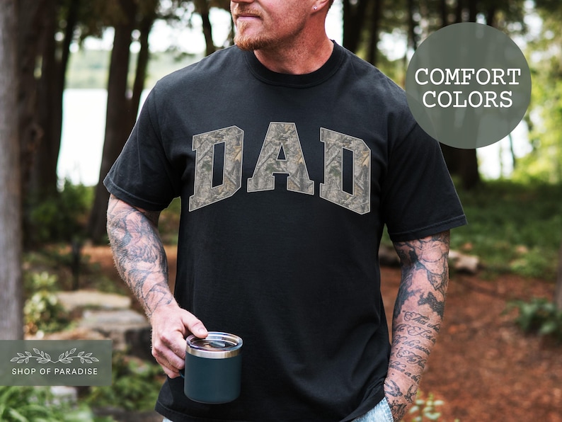 Puede incluir: Camiseta negra con la palabra "DAD" en un dise&ntilde;o de camuflaje en el pecho. La camiseta es de un color c&oacute;modo. El logotipo de Shop of Paradise est&aacute; en la esquina inferior izquierda. La persona sostiene una taza.