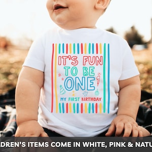 May include: A white t-shirt with the text "IT'S FUN TO BE ONE" in colorful block letters, with the words "MY FIRST BIRTHDAY" below. The shirt has a festive design with vertical stripes and celebratory icons. The shirt is for a child.