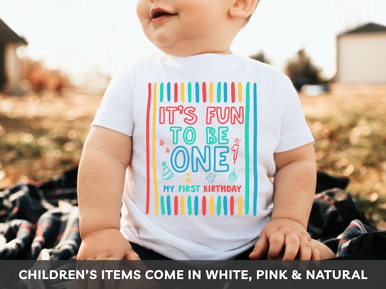 May include: A white t-shirt with the text "IT'S FUN TO BE ONE" in colorful block letters, with the words "MY FIRST BIRTHDAY" below. The shirt has a festive design with vertical stripes and celebratory icons. The shirt is for a child.