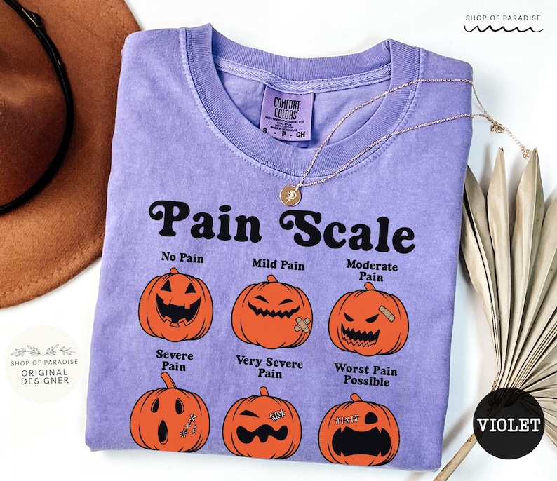 May include: A violet short-sleeved t-shirt featuring a black graphic titled Pain Scale. The design displays six orange pumpkins with varying facial expressions and labels: No Pain, Mild Pain, Moderate Pain, Severe Pain, Very Severe Pain, and Worst Pain Possible.