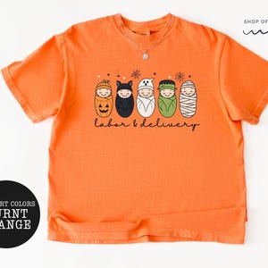 K&ouml;nnte beinhalten: Ein kurz&auml;rmeliges T-Shirt in gebranntem Orange mit einer Grafik von f&uuml;nf in Windeln gewickelten Babys, die als K&uuml;rbis, Fledermaus, Geist, Frankenstein und Mumie verkleidet sind. Der Text "labor & delivery" steht darunter, "SHOP OF PARADISE" oben.