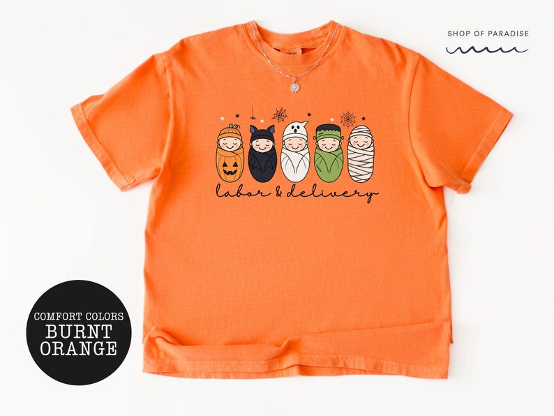 K&ouml;nnte beinhalten: Ein kurz&auml;rmeliges T-Shirt in gebranntem Orange mit einer Grafik von f&uuml;nf in Windeln gewickelten Babys, die als K&uuml;rbis, Fledermaus, Geist, Frankenstein und Mumie verkleidet sind. Der Text "labor & delivery" steht darunter, "SHOP OF PARADISE" oben.