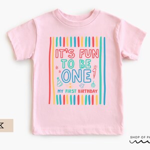 May include: Pink short-sleeved t-shirt with the text "IT'S FUN TO BE ONE" in colorful letters. The shirt has vertical stripes in red, yellow, blue, and green. Below the text is "MY FIRST BIRTHDAY".