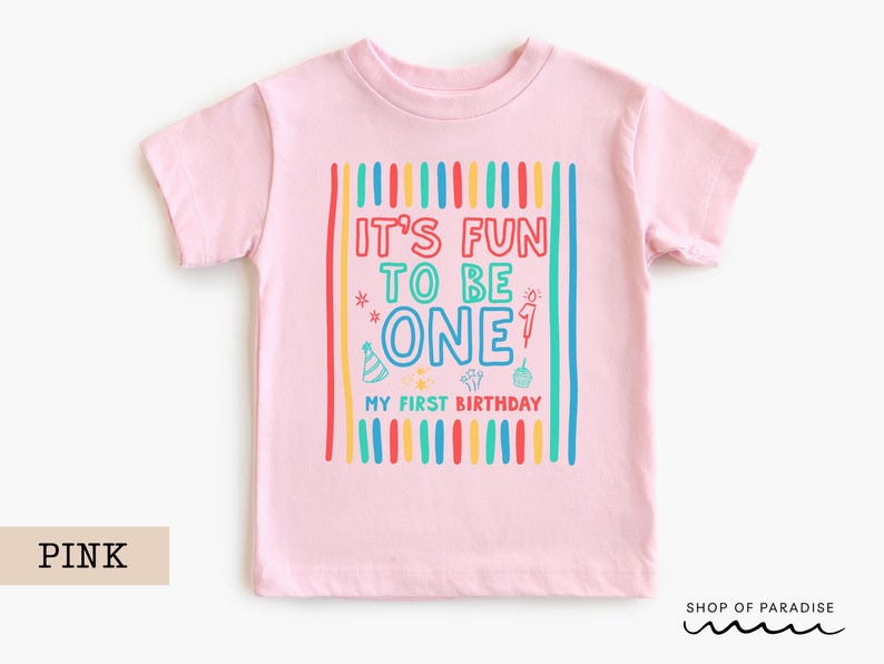 May include: Pink short-sleeved t-shirt with the text "IT'S FUN TO BE ONE" in colorful letters. The shirt has vertical stripes in red, yellow, blue, and green. Below the text is "MY FIRST BIRTHDAY".