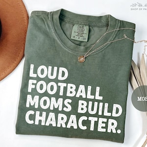 May include: An olive green t-shirt with the text "LOUD FOOTBALL MOMS BUILD CHARACTER" in white block letters. A gold necklace with a pendant rests on the shirt. The shirt is folded and has a tag that says "COMFORT COLORS".