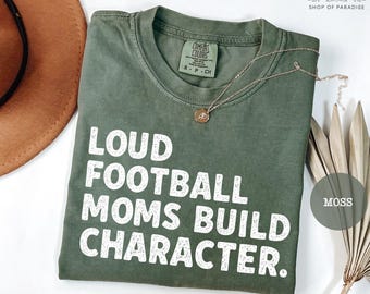 May include: An olive green t-shirt with the text "LOUD FOOTBALL MOMS BUILD CHARACTER" in white block letters. A gold necklace with a pendant rests on the shirt. The shirt is folded and has a tag that says "COMFORT COLORS".