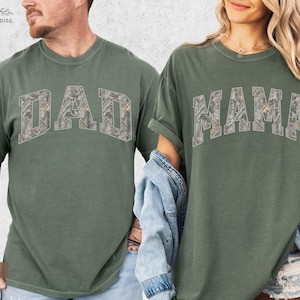 Puede incluir: Dos camisetas verde musgo. Una camiseta tiene la palabra "DAD" en un dise&ntilde;o de camuflaje en el pecho. La otra camiseta tiene la palabra "MAMA" en un dise&ntilde;o de camuflaje en el pecho. Ambas camisetas tienen mangas cortas.