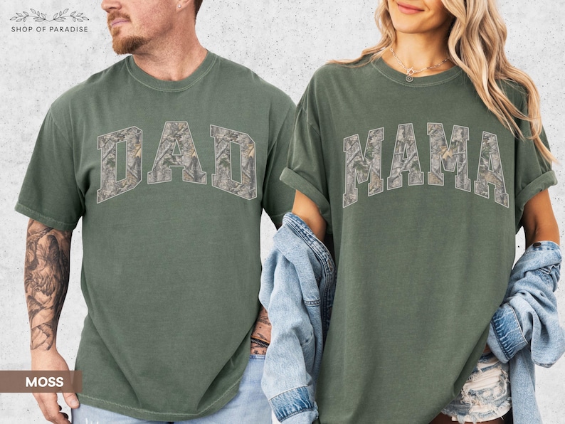 Puede incluir: Dos camisetas verde musgo. Una camiseta tiene la palabra "DAD" en un dise&ntilde;o de camuflaje en el pecho. La otra camiseta tiene la palabra "MAMA" en un dise&ntilde;o de camuflaje en el pecho. Ambas camisetas tienen mangas cortas.