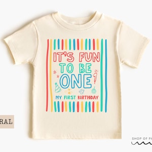 May include: A cream-colored t-shirt with the text "IT'S FUN TO BE ONE" in colorful letters. The shirt has a birthday theme with a party hat, cupcake, and fireworks graphic. The words "MY FIRST BIRTHDAY" are also printed on the shirt.