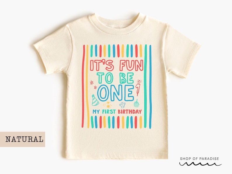 May include: A cream-colored t-shirt with the text "IT'S FUN TO BE ONE" in colorful letters. The shirt has a birthday theme with a party hat, cupcake, and fireworks graphic. The words "MY FIRST BIRTHDAY" are also printed on the shirt.