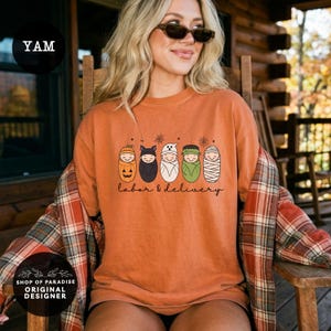 K&ouml;nnte beinhalten: Ein orangefarbenes T-Shirt mit einer Grafik von f&uuml;nf in Windeln gewickelten Babys, die als K&uuml;rbis, Fledermaus, Geist, Frankenstein und Mumie verkleidet sind. Der Text unter den Babys lautet labor & delivery in schwarzer Schreibschrift.