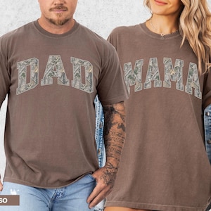 Puede incluir: Dos camisetas color espresso, una con "DAD" y otra con "MAMA" en un dise&ntilde;o de camuflaje. Las camisetas tienen mangas cortas y un corte relajado. La imagen muestra las camisetas siendo usadas por dos personas.