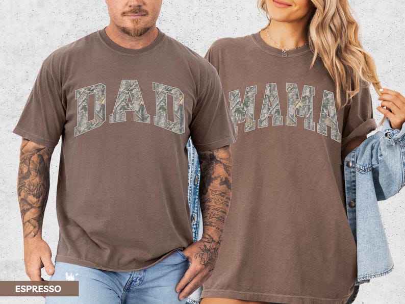 Puede incluir: Dos camisetas color espresso, una con "DAD" y otra con "MAMA" en un dise&ntilde;o de camuflaje. Las camisetas tienen mangas cortas y un corte relajado. La imagen muestra las camisetas siendo usadas por dos personas.