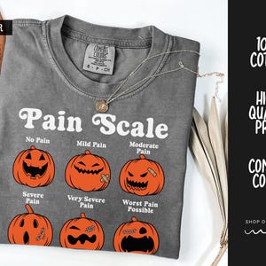 May include: A charcoal gray t-shirt featuring a white graphic titled Pain Scale with six orange jack-o'-lanterns displaying different facial expressions. The shirt has a crew neckline and a Comfort Colors brand label on the inside collar.