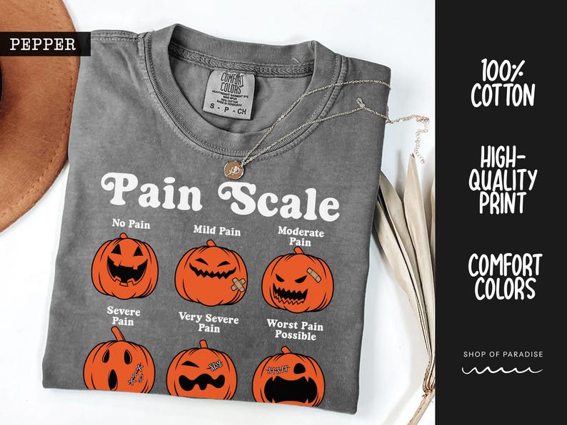 May include: A charcoal gray t-shirt featuring a white graphic titled Pain Scale with six orange jack-o'-lanterns displaying different facial expressions. The shirt has a crew neckline and a Comfort Colors brand label on the inside collar.