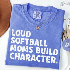 May include: A blue t-shirt with the text "LOUD SOFTBALL MOMS BUILD CHARACTER" in white. The shirt has a crew neck and short sleeves. A gold necklace with a pendant is draped across the shirt. The shirt is labeled "FLO BLUE".