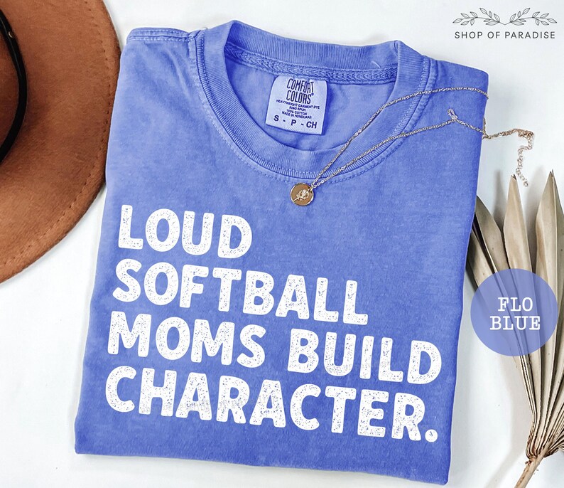 May include: A blue t-shirt with the text "LOUD SOFTBALL MOMS BUILD CHARACTER" in white. The shirt has a crew neck and short sleeves. A gold necklace with a pendant is draped across the shirt. The shirt is labeled "FLO BLUE".