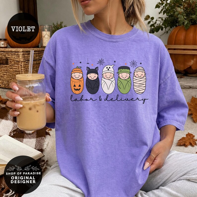 K&ouml;nnte beinhalten: Ein hellviolettes Kurzarm-T-Shirt mit einer Grafik von f&uuml;nf in Windeln gewickelten Babys, die als K&uuml;rbis, Fledermaus, Geist, Frankenstein und Mumie verkleidet sind. Unter den Babys steht der Text labor & delivery in schwarzer Schreibschrift.