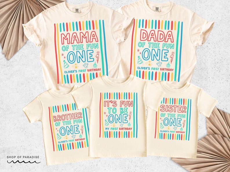 May include: Cream-colored t-shirts with colorful text and graphics. The shirts read "MAMA," "DADA," "BROTHER," "IT'S FUN TO BE," and "SISTER" with "ONE" and "Oliver's First Birthday." The shirts have vertical stripes.