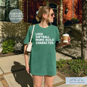 May include: Light green t-shirt with the text "LOUD SOFTBALL MOMS BUILD CHARACTER" in white. The shirt is a Comfort Colors brand. The image also includes a white purse and a coffee cup.