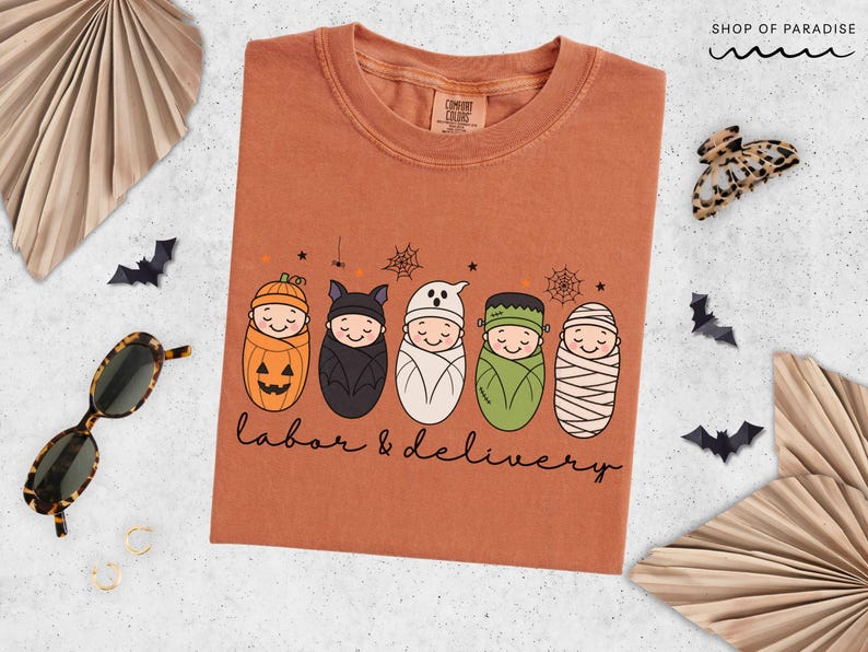 K&ouml;nnte beinhalten: Ein rostfarbenes T-Shirt mit einer Grafik von f&uuml;nf in Windeln gewickelten Babys, die als K&uuml;rbis, Fledermaus, Geist, Frankensteins Monster und Mumie verkleidet sind. Der Text "labor & delivery" steht in schwarzer Schreibschrift unter den Figuren.