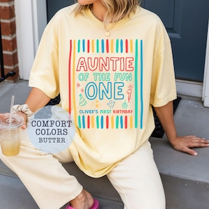 May include: Butter yellow t-shirt with the text "AUNTIE OF THE FUN ONE" in a colorful design. The shirt also includes "Oliver's First Birthday". The shirt is part of the "Comfort Colors" collection.