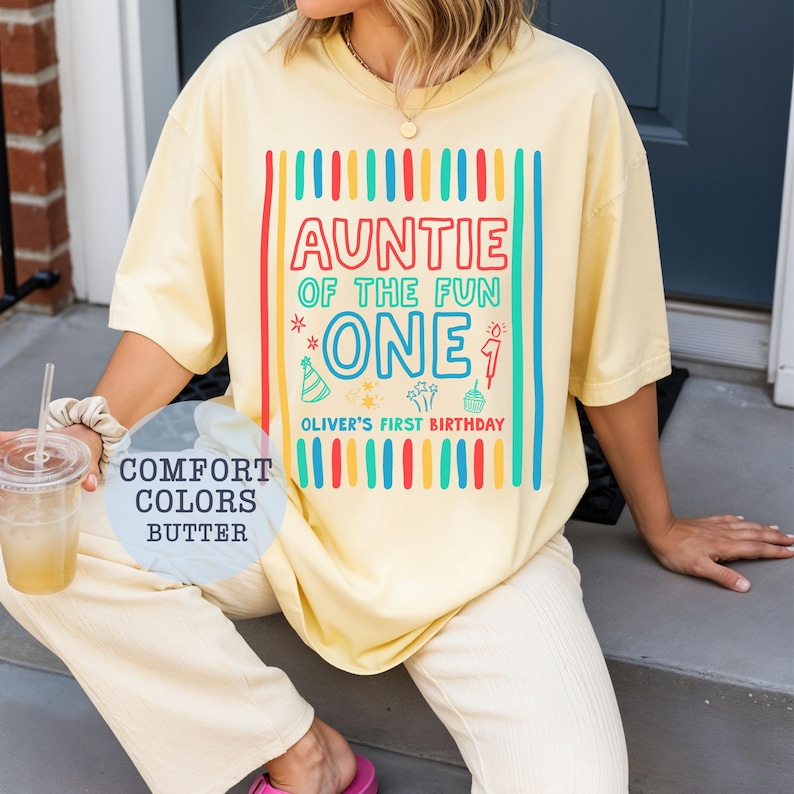 May include: Butter yellow t-shirt with the text "AUNTIE OF THE FUN ONE" in a colorful design. The shirt also includes "Oliver's First Birthday". The shirt is part of the "Comfort Colors" collection.