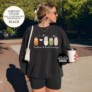 K&ouml;nnte beinhalten: Ein schwarzes Langarm-Sweatshirt mit einer Grafik von f&uuml;nf Babys in Halloween-Kost&uuml;men, darunter ein K&uuml;rbis, eine Fledermaus, ein Geist, Frankenstein und eine Mumie. Der Text "labor & delivery" ist in wei&szlig;er Schreibschrift unter den Babys gedruckt.