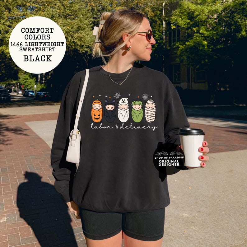 K&ouml;nnte beinhalten: Ein schwarzes Langarm-Sweatshirt mit einer Grafik von f&uuml;nf Babys in Halloween-Kost&uuml;men, darunter ein K&uuml;rbis, eine Fledermaus, ein Geist, Frankenstein und eine Mumie. Der Text "labor & delivery" ist in wei&szlig;er Schreibschrift unter den Babys gedruckt.