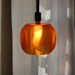 Könnte beinhalten: Eine orangefarbene, würfelförmige Pendelleuchte mit abgerundeten Aussparungen, die an einem schwarzen Arm hängt. Warmes Licht scheint von innen. Im Hintergrund ein weißer Vorhang.