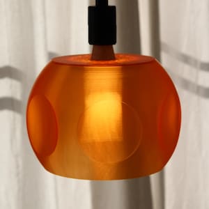 Könnte beinhalten: Orangefarbene Pendelleuchte mit kugelförmigem Schirm. Der Schirm hat drei kreisförmige Aussparungen und einen schwarzen Stiel. Das Licht ist eingeschaltet und erzeugt einen warmen Schein. Die Leuchte hängt vor einem weißen Vorhang.