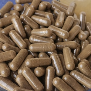May include: A close-up of many brown capsules, possibly supplements or medications, filling the image. The capsules are cylindrical with rounded ends, and the contents are a light brown powder. The capsules are in a clear container.