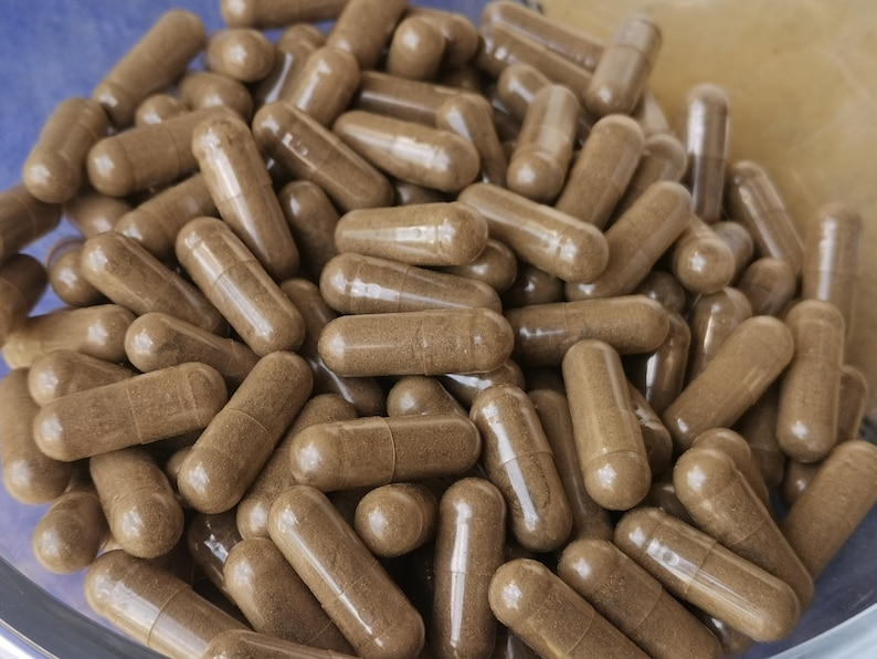 May include: A close-up of many brown capsules, possibly supplements or medications, filling the image. The capsules are cylindrical with rounded ends, and the contents are a light brown powder. The capsules are in a clear container.
