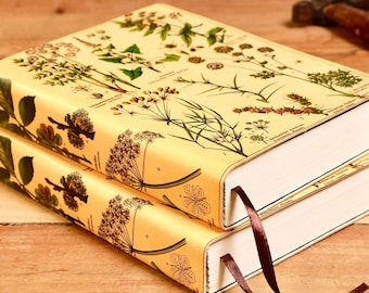 Puede incluir: Dos libros de tapa dura apilados sobre una superficie de madera. Las cubiertas son de color amarillo claro con ilustraciones botánicas y texto. Cada libro tiene una cinta de marcador marrón. Las páginas son blancas.