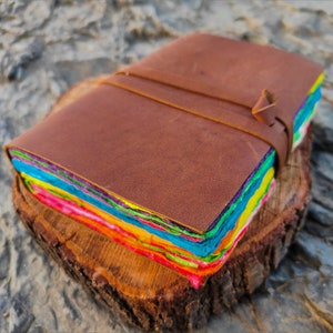 Op de afbeelding: Een bruin lederen gebonden dagboek met een regenboogkleurige stapel papier erin. Het dagboek is gesloten en ligt op een houten oppervlak.