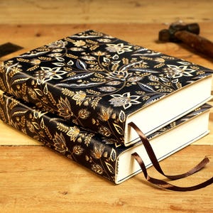 May include: Two black leather-bound journals with a gold and blue floral pattern on the cover. The journals are stacked on top of each other and have brown ribbon bookmarks.