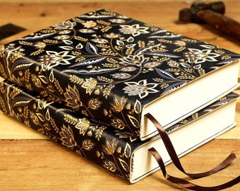 May include: Two black leather-bound journals with a gold and blue floral pattern on the cover. The journals are stacked on top of each other and have brown ribbon bookmarks.