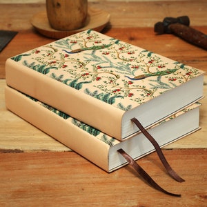 Może przedstawiać: Dwa kremowe dzienniki z wzorem pawia i kwiatów na okładce. Dzienniki mają brązowe wstążki jako zakładki i są ułożone jeden na drugim.