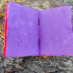 May include: A purple handmade journal with a red cover and brown leather binding. The journal is open to reveal blank pages.